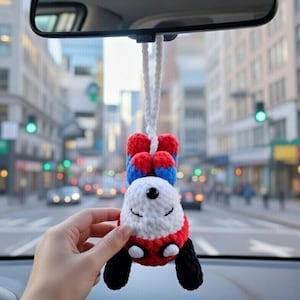 May include: A handmade crochet car charm in the shape of a smiling panda. The panda has a white face, red and black accents, and is suspended by a white cord. The charm is hanging from a car's rearview mirror.