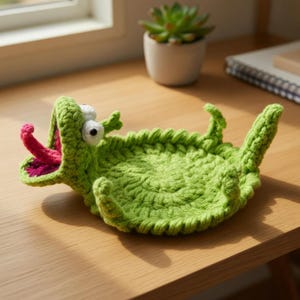 May include: A handmade, green crochet alligator-shaped tray with white eyes and a pink tongue. The tray is lying on a wooden surface. The alligator's mouth is open, and its tongue is visible. The tray is designed to hold small items.