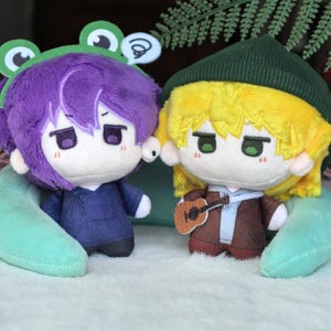 Pu&ograve; includere: Due bambole di peluche con caratteristiche in stile anime. Una bambola ha i capelli viola e un cappello da rana, l'altra ha i capelli gialli e un berretto verde. La bambola dai capelli gialli tiene in mano una chitarra in miniatura. Entrambe le bambole hanno espressioni tristi.