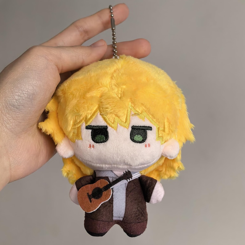 Pu&ograve; includere: Bambola di peluche con capelli gialli, occhi verdi e un abito marrone, che tiene una piccola chitarra. La bambola ha un portachiavi attaccato. Il viso della bambola presenta dettagli ricamati, tra cui piccoli segni rossi.