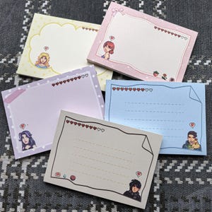 May include: A collection of five colorful sticky note pads. Each pad features a different pastel color, decorative borders, and illustrations of characters. The pads have heart-shaped accents and lined spaces for writing.
