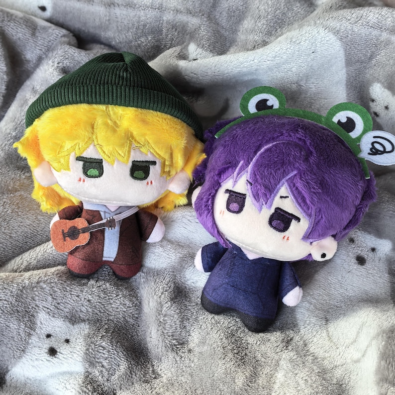 Pu&ograve; includere: Due bambole di peluche con caratteristiche in stile anime. Una bambola ha i capelli gialli, un berretto verde e una piccola chitarra. L'altra bambola ha i capelli viola e una fascia a tema rana. Entrambe le bambole hanno gli occhi verdi e sono su una coperta grigia.