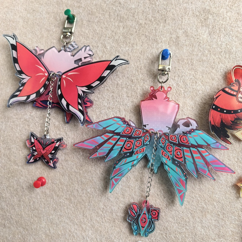 May include: Two acrylic butterfly charms with chain attachments. One charm features red and black wings, the other has teal and red wings. Both have a small charm dangling below. Each charm has a silver-colored clasp.