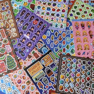May include: A collection of colorful sticker sheets featuring pixelated art. The stickers depict characters, objects, and scenes from a video game. Sheets include images of people, food, and other game-related items. The stickers are in various shapes and sizes.