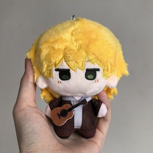 Pu&ograve; includere: Bambola di peluche con capelli gialli, occhi verdi e una piccola chitarra marrone. La bambola indossa una giacca marrone sopra una camicia bianca e una cravatta grigia. La bambola &egrave; un portachiavi.