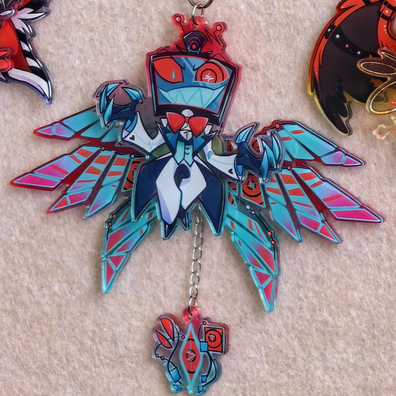 May include: A colorful keychain featuring a stylized character with a box-shaped head, a red bow tie, and blue wings with red and pink accents. The character is wearing a tuxedo. A smaller charm hangs below, connected by a chain.