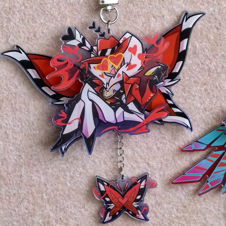 May include: A clear acrylic keychain featuring a stylized character design with red, black, white, and yellow accents. The keychain has a butterfly-like shape with a smaller, matching charm hanging below. A silver-colored clasp is at the top.