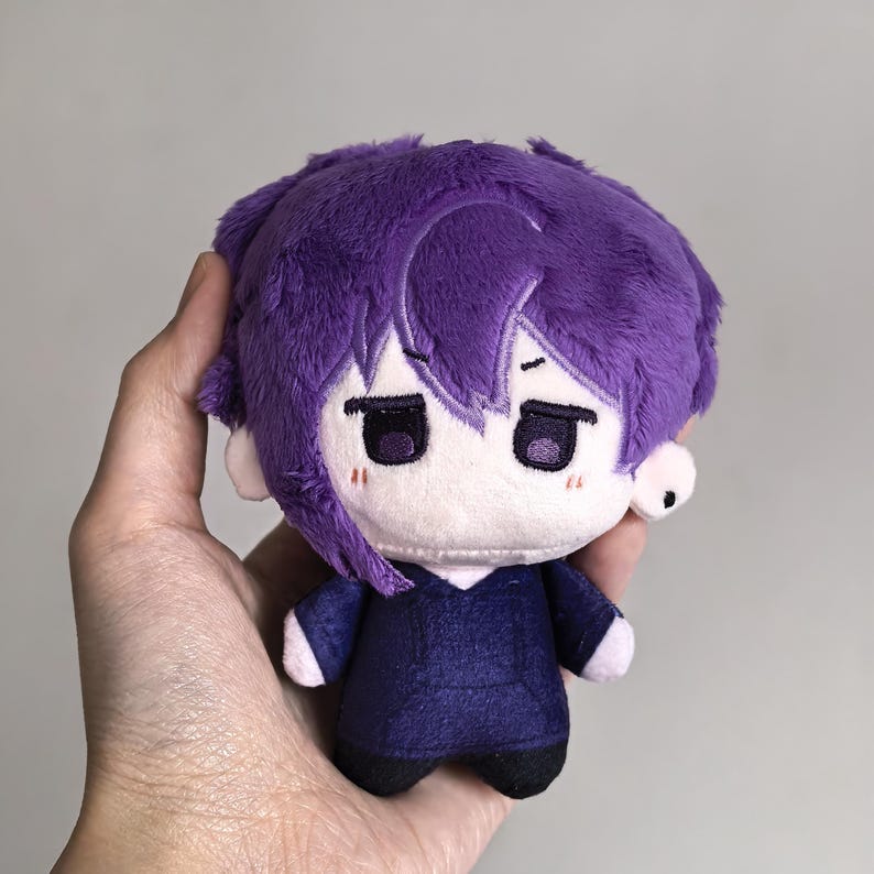 Pu&ograve; includere: Una bambola di peluche dal design in stile cartone animato. La bambola ha i capelli viola, un viso bianco e un completo viola scuro. La bambola &egrave; tenuta in mano su uno sfondo neutro.
