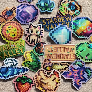 May include: A collection of pixelated stickers featuring various designs and the text "Stardew Valley." The stickers showcase colorful graphics of hearts, gems, and other game-related elements. The stickers are arranged on a neutral-colored surface.