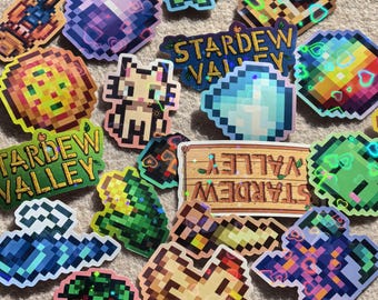 Puede incluir: Una colección de pegatinas pixeladas con varios diseños y el texto "Stardew Valley". Las pegatinas muestran gráficos coloridos de corazones, gemas y otros elementos relacionados con el juego. Las pegatinas están dispuestas sobre una superficie de color neutro.