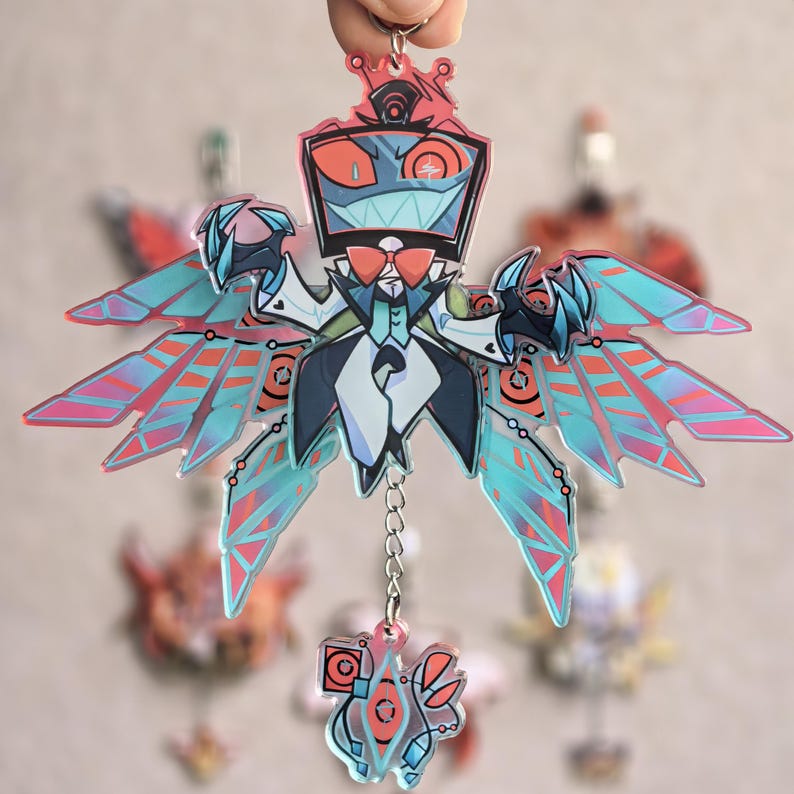 May include: A clear acrylic keychain featuring a stylized robot character with a crown, bow tie, and wings. The robot has a red and teal color scheme with a small charm hanging below. Other keychains are blurred in the background.