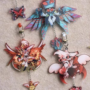 May include: A collection of colorful acrylic keychains featuring various cartoon characters with wings and intricate designs. The keychains are attached to metal clasps and chains. The color palette includes red, blue, and orange.