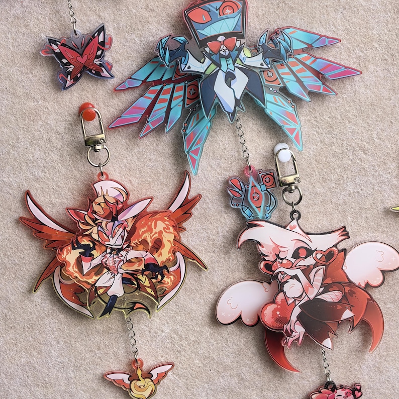 May include: A collection of colorful acrylic keychains featuring various cartoon characters with wings and intricate designs. The keychains are attached to metal clasps and chains. The color palette includes red, blue, and orange.