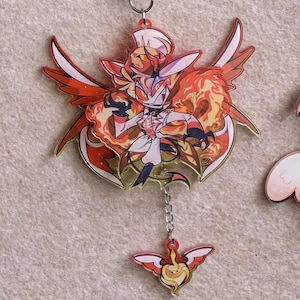 May include: A red and gold acrylic keychain featuring a stylized character with fiery wings and a crown. The character is wearing a white and black outfit. A smaller charm with a flame design hangs below.