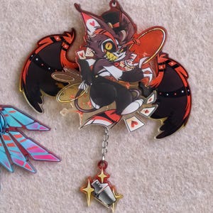 May include: A clear acrylic charm featuring a cartoon character with a cat-like face, playing cards. The character has red, black, and white accents, with large wings and a top hat. A small charm of a cocktail shaker and stars hangs below.