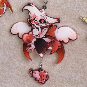 May include: A red and white acrylic charm featuring a cartoon character with heart-shaped glasses and wings. The charm has a small pink elephant attached by a chain. The character is in a sitting pose, with a dark red cape and black fishnet stockings.