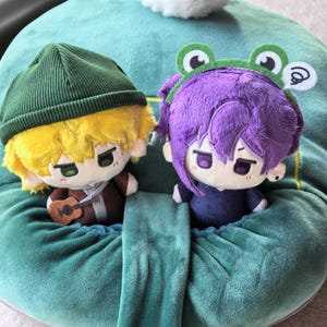 Pu&ograve; includere: Due bambole di peluche con caratteristiche in stile anime. Una bambola ha i capelli gialli e un berretto verde, che tiene una piccola chitarra. L'altra bambola ha i capelli viola e una fascia a tema rana. Entrambe le bambole hanno tratti del viso dettagliati.