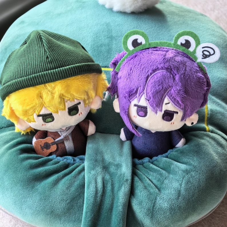 Pu&ograve; includere: Due bambole di peluche con caratteristiche in stile anime. Una bambola ha i capelli gialli e un berretto verde, che tiene una piccola chitarra. L'altra bambola ha i capelli viola e una fascia a tema rana. Entrambe le bambole hanno tratti del viso dettagliati.