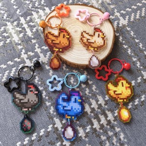 May include: A selection of pixelated chicken keyrings in shades of brown, blue, and yellow. Each keyring showcases a chicken design with a dangling egg-shaped charm. The keyrings have different coloured star and flower-shaped accents.