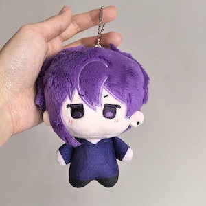 Pu&ograve; includere: Una bambola di peluche con capelli viola e un abito abbinato. La bambola ha un piccolo portachiavi argentato attaccato alla sommit&agrave; della testa. Il viso della bambola ha un'espressione neutra con occhi scuri e un piccolo rossore su ogni guancia.