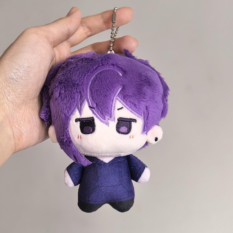 Pu&ograve; includere: Una bambola di peluche con capelli viola e un abito abbinato. La bambola ha un piccolo portachiavi argentato attaccato alla sommit&agrave; della testa. Il viso della bambola ha un'espressione neutra con occhi scuri e un piccolo rossore su ogni guancia.
