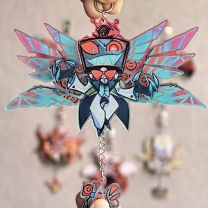 May include: A colorful acrylic keychain featuring a cartoon character with a red bow tie, a black top hat, and large, stylized wings with pink and blue accents. The keychain is attached to a silver keyring and chain.