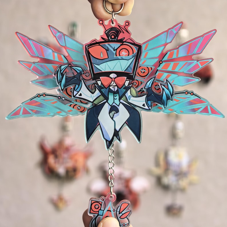 May include: A colorful acrylic keychain featuring a cartoon character with a red bow tie, a black top hat, and large, stylized wings with pink and blue accents. The keychain is attached to a silver keyring and chain.