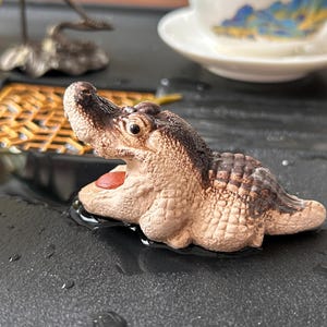 May include: A small, detailed figurine of an alligator with its mouth open, revealing a red interior. The alligator is primarily tan with darker brown accents and sits in a shallow pool of water. The figurine is likely ceramic or a similar material.