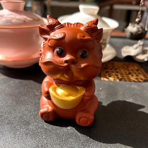 May include: A reddish-brown figurine of a mythical creature, possibly a dragon or a lion, holding a gold ingot. The figurine has large black eyes and small horns. The background includes a pink tea cup and a white tea pot.