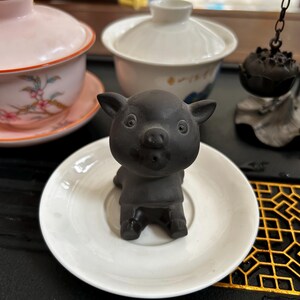 Peut inclure: Une figurine de petit cochon en céramique noire posée sur une soucoupe blanche. Le cochon est entouré d'autres éléments de cérémonie du thé, dont une tasse rose à motifs floraux et un bol blanc avec couvercle.