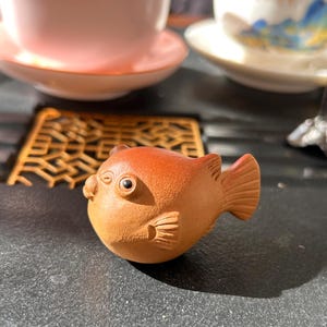 Peut inclure: Petite figurine de poisson-globe en céramique brun-orangé. Le poisson a des nageoires détaillées et un corps rond avec une expression légèrement renfrognée. Il s'agit probablement d'un objet décoratif ou d'un animal de compagnie pour le thé.