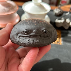 May include: A dark brown, ceramic tea pet in the shape of a smiling frog. The tea pet has detailed facial features and a textured surface. Other tea ceremony items are visible in the background.