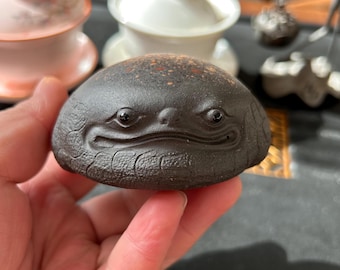 May include: A dark brown ceramic tea pet, shaped like a smiling frog. The tea pet has detailed facial features and a textured surface. Other tea ceremony items are visible in the background.
