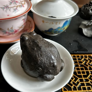 May include: A dark grey frog ornament on a small white plate. The frog has a textured surface, and is surrounded by teacups with floral and landscape designs. The scene suggests a tea ceremony setting.