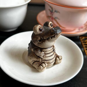 Puede incluir: Pequeña figura de cerámica de un caimán con ojos grandes y redondos y un cuerpo detallado y texturizado. El caimán es marrón y beige, sentado sobre un platillo blanco. Otras tazas de té son visibles en el fondo.