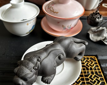 May include: A black ceramic figurine of a mythical creature, possibly a Foo Dog, rests on a white saucer. A pink teacup with floral designs and a white teacup with a lid are also visible. A tea infuser hangs nearby.