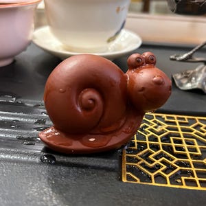 May include: A whimsical, reddish-brown ceramic snail figurine with a smiling face and a spiraled shell. The snail is positioned on a dark surface with a gold-colored geometric pattern.