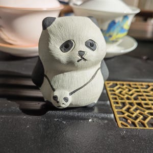 May include: A ceramic panda figurine with black and white details. The panda has a small baby panda attached to its belly. The figurine has a sad expression and is sitting on a dark surface. Tea cups are in the background.