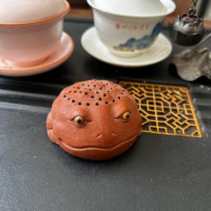 以下が含まれることがあります： 黄色い目と頭に多数の小さな穴がある、赤茶色のセラミック製のカエルの茶寵。 カエルはティーセットの一部で、ピンクのティーカップと、背景に青と金の風景デザインが施された白いティーカップがあります。