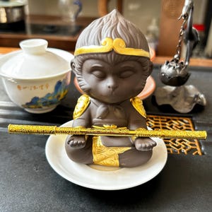 Puede incluir: Una figura de cerámica marrón de un mono sentado, sosteniendo un bastón dorado. El mono tiene detalles dorados en la cabeza y los hombros. Está sobre un plato blanco. Una taza de té blanca con detalles pintados en azul y dorado está en el fondo.