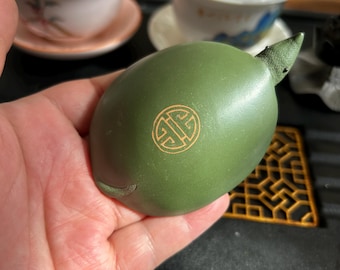 May include: A green ceramic tea pet shaped like a lemon, featuring a small stem and a gold emblem. The tea pet is held in a hand, with teacups in the background. This item is used in tea ceremonies.