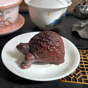 Puede incluir: Figurita detallada de tortuga marrón rojizo con caparazón texturizado, sobre un plato blanco pequeño. La tortuga está tallada con detalles intrincados, mostrando su cabeza, patas y patrón de caparazón. Accesorios de té.