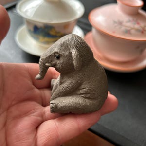 May include: A small, detailed figurine of a sitting elephant in a grey-brown colour. The elephant has a textured surface and is resting on a hand. In the background, there are two tea cups with lids.