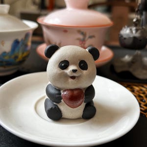 Puede incluir: Una figura de panda de cerámica con orejas, ojos y extremidades negras, sosteniendo un corazón rojo. El panda está sentado en un platillo blanco, con un fondo borroso con tazas de té y un juego de té rosa.