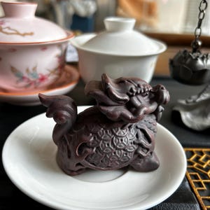 May include: A detailed, dark brown dragon figurine rests on a small white saucer. The dragon is intricately carved with scales and a detailed mane. In the background, there are two teacups and a tea strainer, suggesting a tea-related theme.