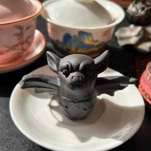 Könnte beinhalten: Eine dunkelgraue Fledermausfigur mit ausgebreiteten Flügeln sitzt auf einer kleinen weißen Untertasse. Die Fledermaus hat detaillierte Merkmale, einschließlich spitzer Ohren und sichtbarer Fangzähne. Weitere Tee-Artikel sind im Hintergrund.