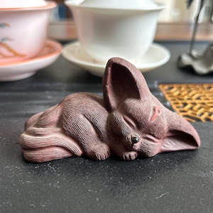 以下が含まれることがあります： 眠っているポーズの、詳細な赤茶色のキツネの置物。 キツネは目を閉じ、小さな黒い目と質感のある表面を持っています。 この置物は、おそらくセラミックまたは同様の素材で作られています。 背景にはティーカップがあります。