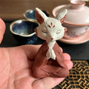 May include: A small ceramic fox figurine with a white head and ears, featuring red markings on the forehead and ears. The fox has closed eyes and is depicted in a seated, meditative pose with paws pressed together, wearing a dark brown robe.