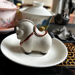 May include: A small, off-white ceramic horse figurine with a red saddle and black hooves. The horse is standing on a white saucer. A small gold-colored object sits on the horse's back. Other ceramic tea sets are in the background.