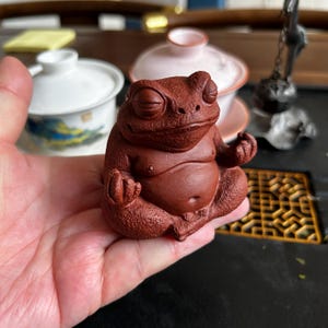 Könnte beinhalten: Eine rotbraune Keramik-Froschfigur in einer sitzenden Meditationspose. Der Frosch hat geschlossene Augen und einen ruhigen Ausdruck. Die Figur wird in einer Hand gehalten, mit anderen Teezeremonie-Artikeln im Hintergrund.